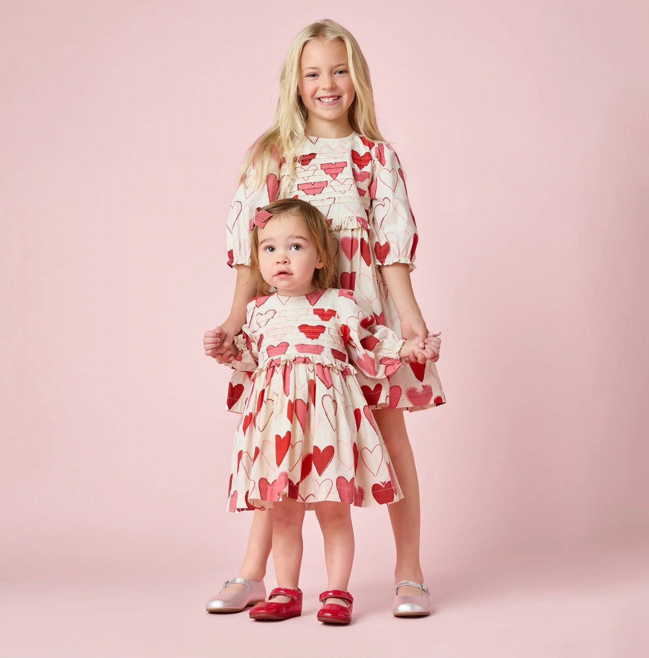 Two young girls pose before a pink background, both in cream dresses. The younger wears Pink Chicken’s Gradient Hearts Renee Dress, featuring heart prints, a pleated bodice, and a red bow. The older girl smiles with her long blonde hair.