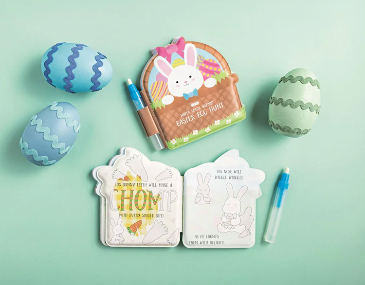 The mudpie Easter Water Color Wizard Books set includes a bunny-shaped book with unfinished bunny illustrations, two water pens for mess-free painting, two decorated blue eggs, and one green egg, all displayed on a light green surface.