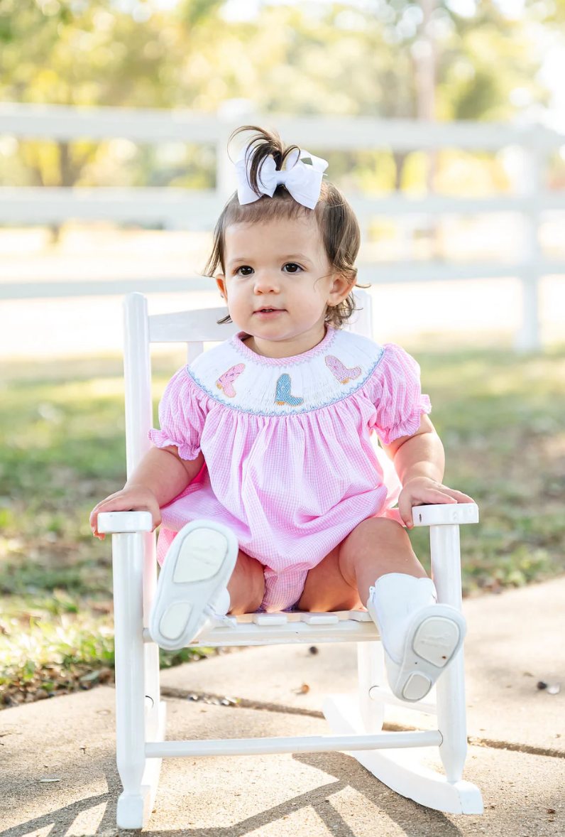 A toddler with brown hair and a white bow sits on a small white rocking chair outside, wearing the Ruth & Ralph Cowgirl Boots Smocked Birdie Bubble. The scene features green grass, trees, and a white fence in the background.