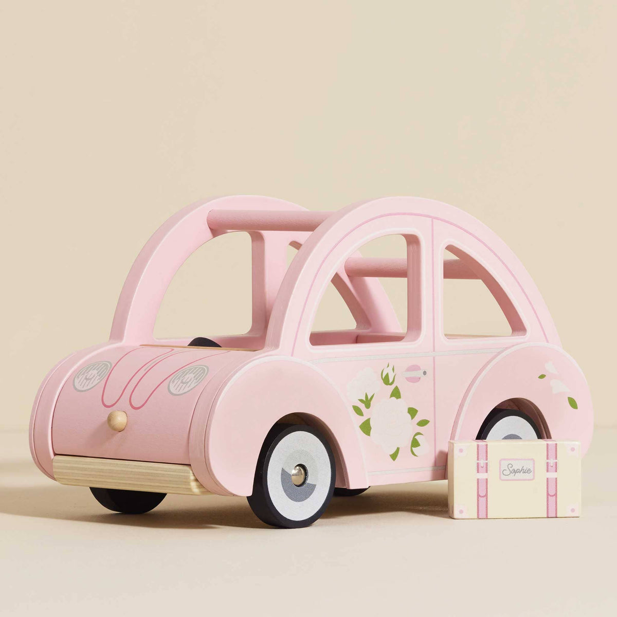The Sophie's Dolls House Toy Car by Le Toy Van, Inc. is a retro pink wooden car with floral details, shown beside a matching pink and white suitcase labeled Sophie—a charming, plastic-free toy on a neutral background.