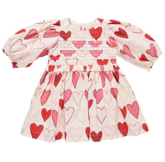 The Gradient Hearts Renee Dress by Pink Chicken is a white long-sleeve children's dress with pleated bodice, gathered waist, puffed sleeves, and allover pink and red heart prints.