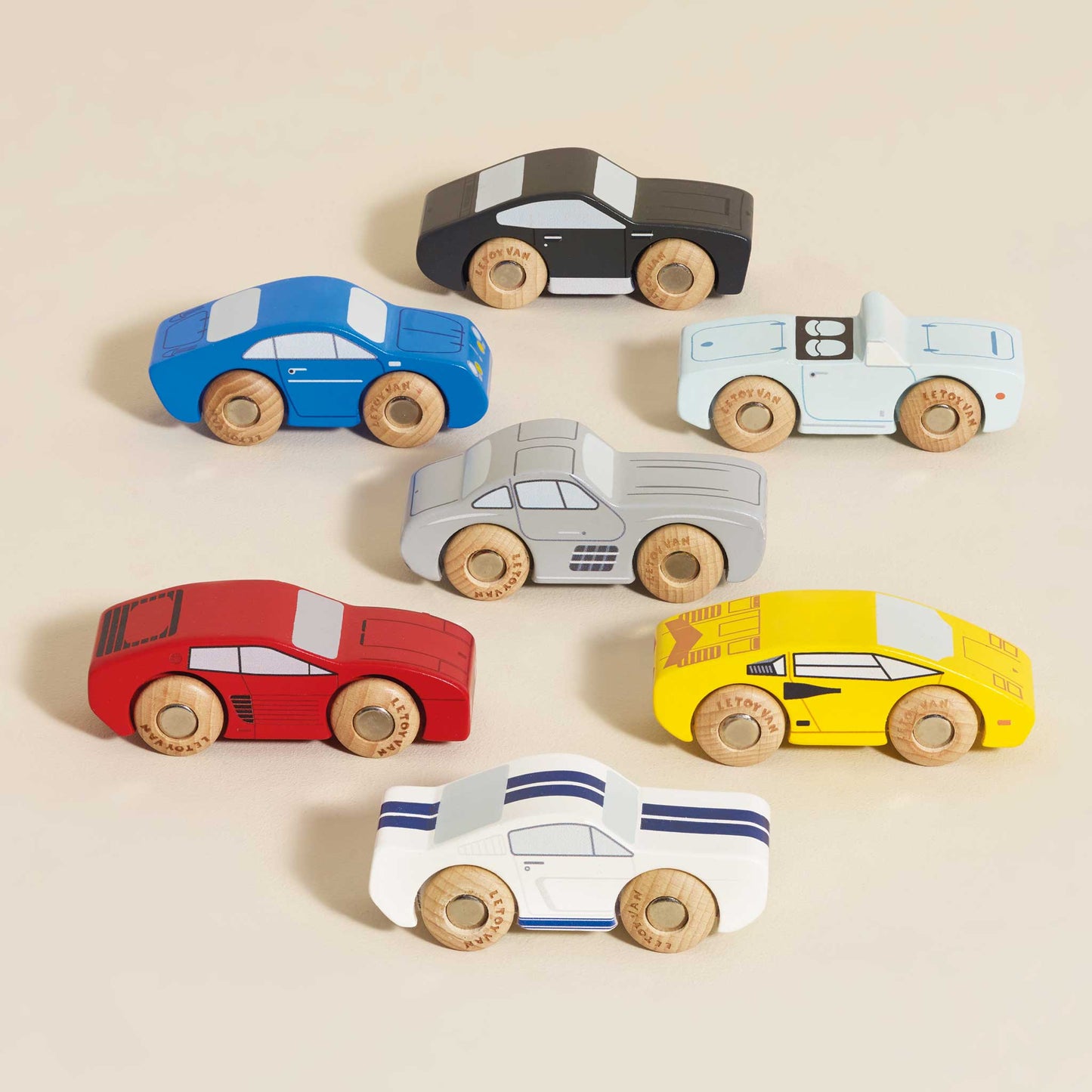 Seven Classic Wooden Sports Cars by Le Toy Van, Inc. in blue, black, light blue, red, grey, yellow, and white with blue stripes are arranged on a beige surface and are compatible with train tracks.