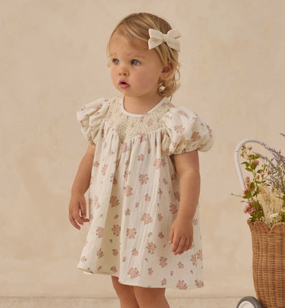 Young girl in a floral dress with a bow, standing next to a basket of flowers against a beige background