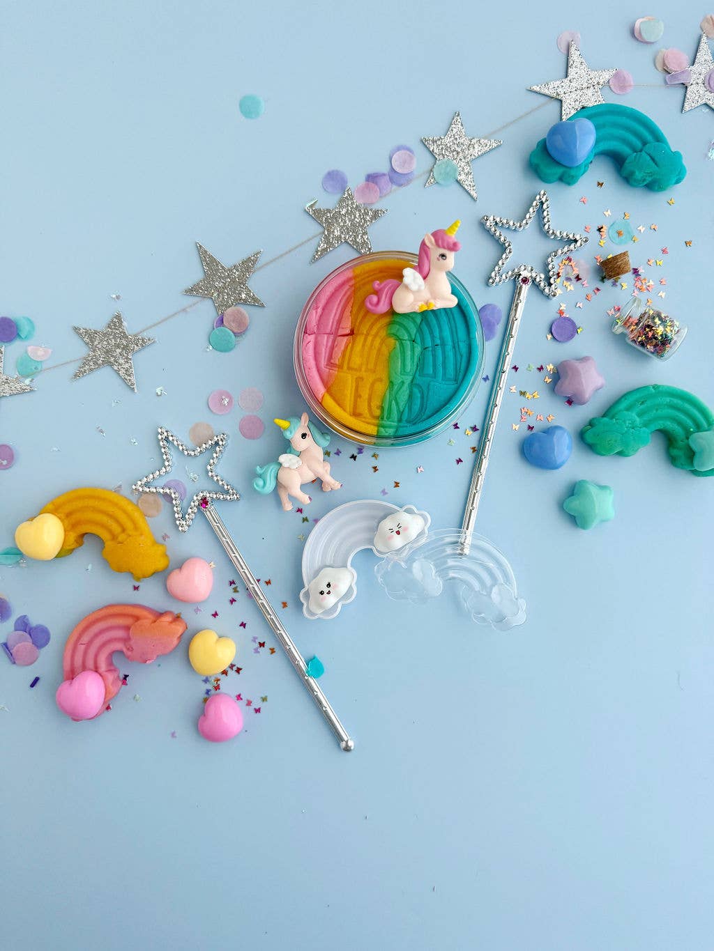 Colorful flat lay with unicorn and rainbow toys, non-toxic modeling compound, silver star wands, confetti, glitter, pastel charms, and the Unicorn KidDough Play Kit by Earth Grown KidDoughs on a light blue background with a silver star garland.