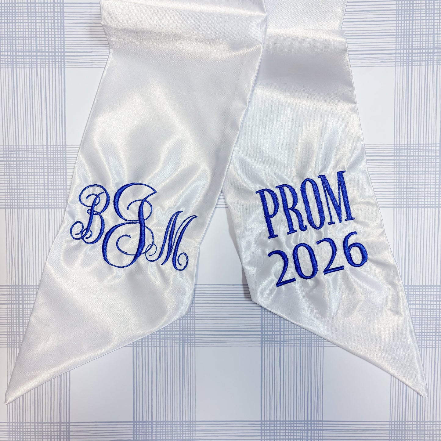 White satin sash with blue Classic monogram  and 'PROM 2026'  text on a checkered background