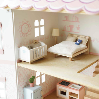 The Complete Dolls House Furniture by Le Toy Van, Inc. includes a miniature bedroom with a bed, white blanket, blue book, crib with teddy bear, lamp, window wall decals—and a kitchen with stove, fridge, plant, and more.