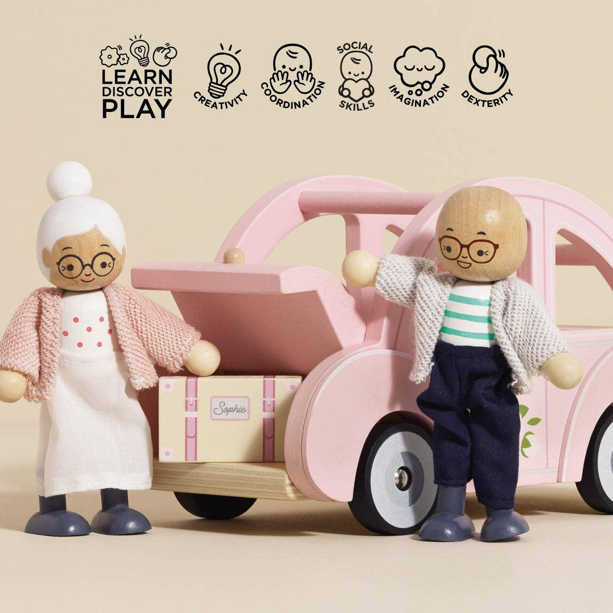 Two wooden dolls (grandmother and child) from Le Toy Van, Inc.'s Sophie's Dolls House Toy Car stand beside a retro car with an open trunk and suitcase. Icons show it boosts creativity, coordination, social skills, imagination, and dexterity—plastic-free fun!.