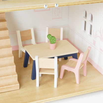 The Complete Dolls House Furniture set by Le Toy Van, Inc. includes a wooden dining table, four colorful chairs, and a potted cactus centerpiece, perfect for furnishing a dollhouse dining room near the stairs with white walls and window cutouts.