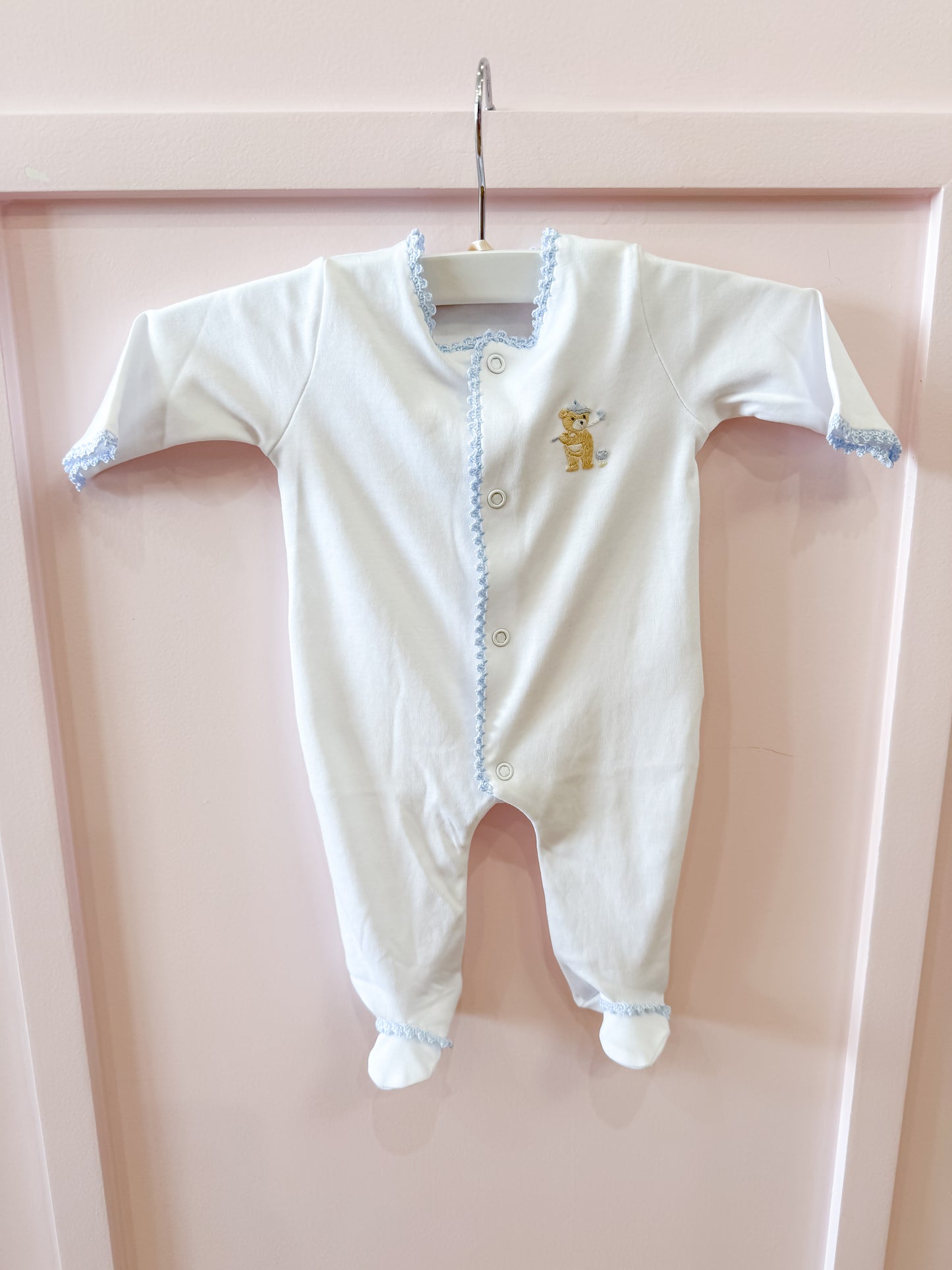 The Baby Club Chic "Golfing Bear Footie" in white with blue trim hangs on a white hanger against a pale pink background. It features long sleeves, covered feet, and an embroidered bear with a blue crown on the chest.