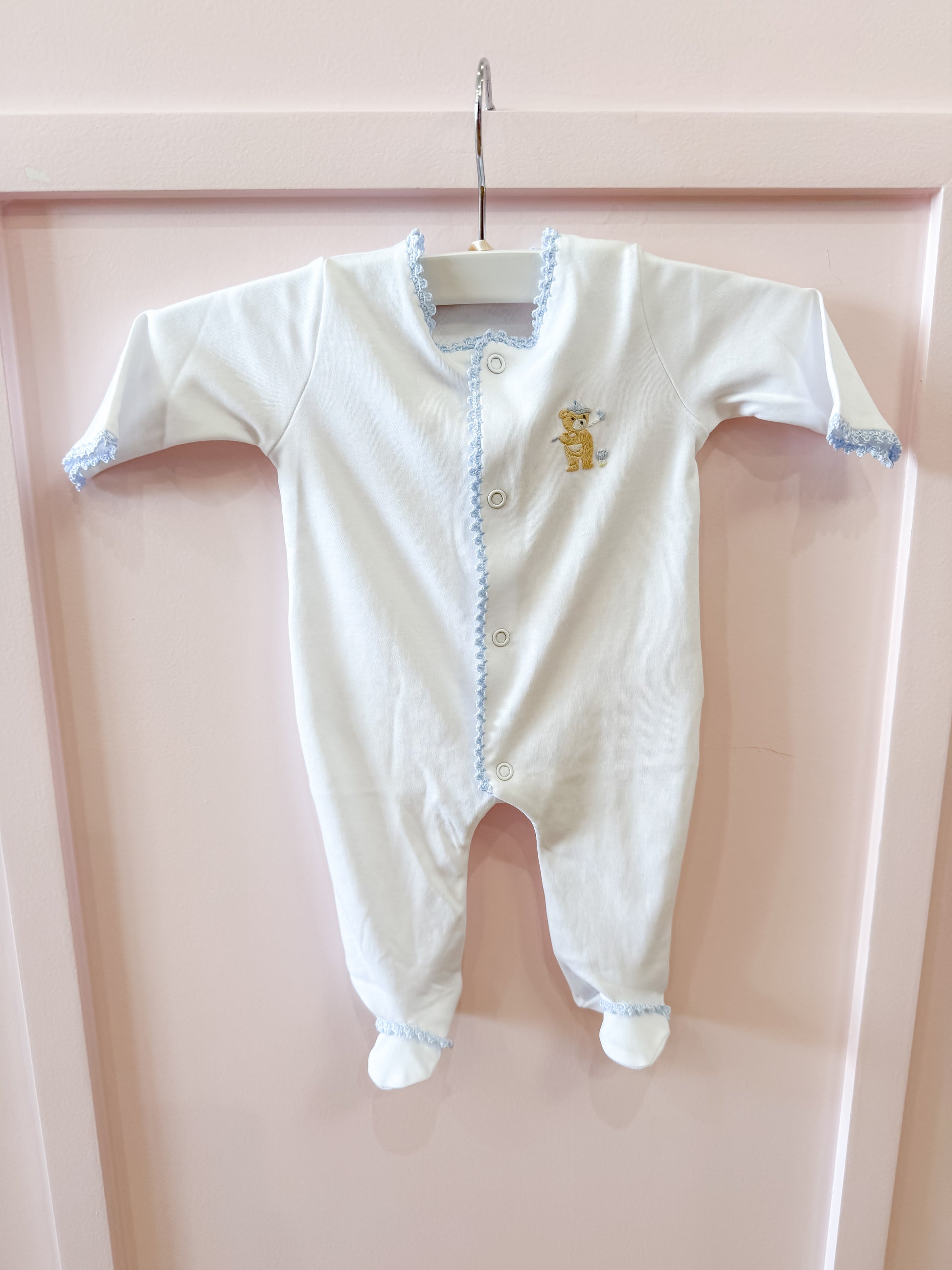 The Baby Club Chic "Golfing Bear Footie" in white with blue trim hangs on a white hanger against a pale pink background. It features long sleeves, covered feet, and an embroidered bear with a blue crown on the chest.