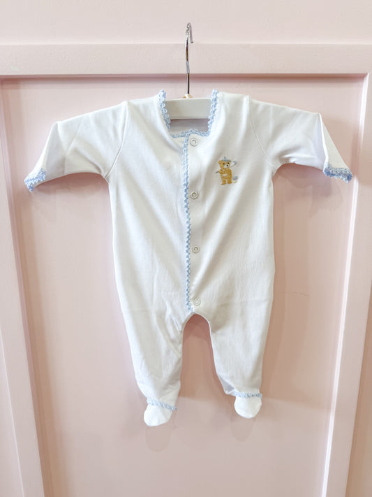 The Baby Club Chic "Golfing Bear Footie" in white with blue trim hangs on a white hanger against a pale pink background. It features long sleeves, covered feet, and an embroidered bear with a blue crown on the chest.