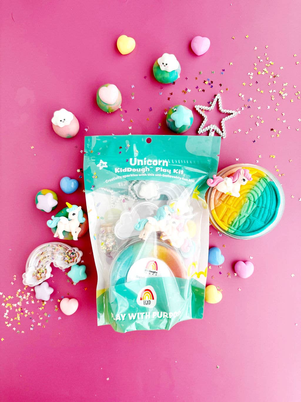 The Unicorn KidDough Play Kit by Earth Grown KidDoughs comes in a teal bag with non-toxic modeling compound, set on a pink background and surrounded by mini rainbow, heart, and star toys plus pastel glitter decorations.