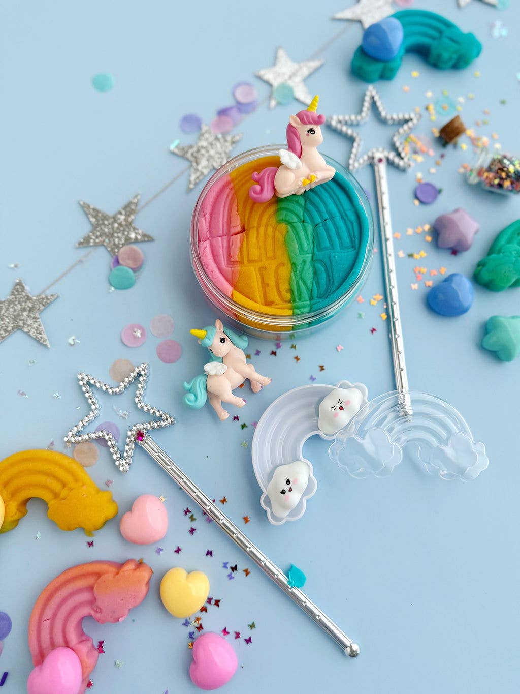 Colorful flat lay on blue features unicorn and rainbow figurines, confetti, hearts, star wands, glittery stars, and the Earth Grown KidDoughs Unicorn KidDough Play Kit with sensory dough in a multicolored circular container at the center.