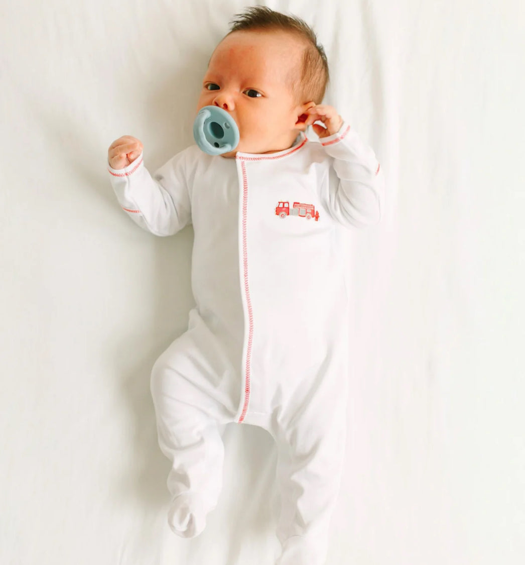 A baby in the Magnolia Baby To the Rescue Footie, featuring a small red fire truck design, lies on a white sheet with a blue pacifier in their mouth.