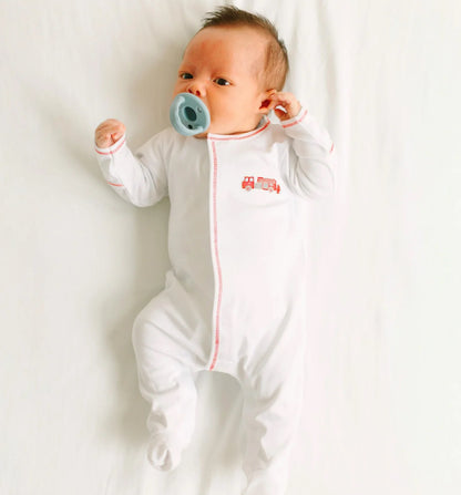 A baby in the Magnolia Baby To the Rescue Footie, featuring a small red fire truck design, lies on a white sheet with a blue pacifier in their mouth.