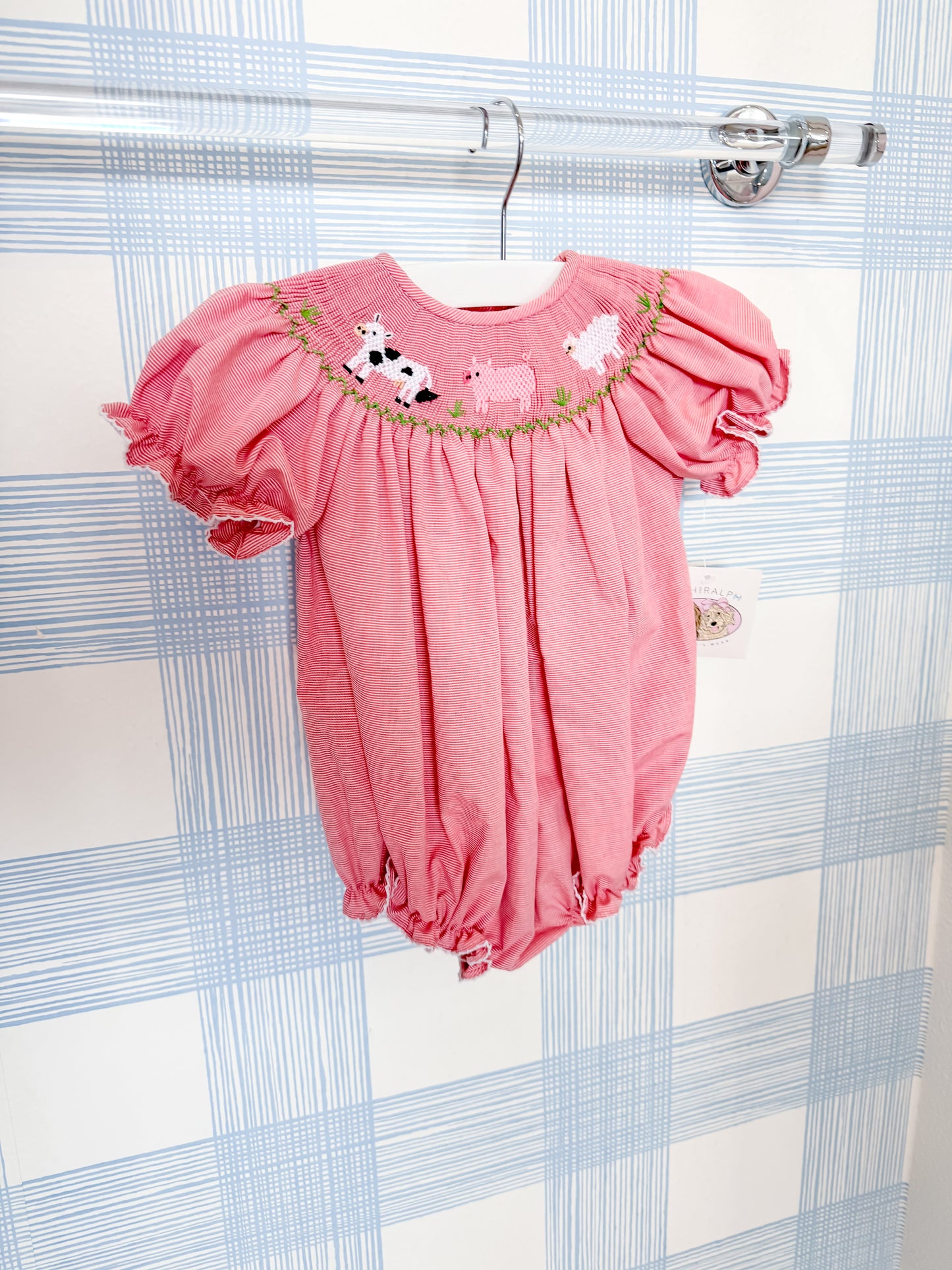 The Ruth & Ralph Farm Animal Bubble, a pink smocked baby romper with puffy sleeves and embroidered cows and pigs, hangs on a clear hanger against a blue and white plaid wall.