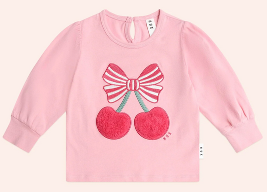 HUX Cherry Pie Puff Sleeve Top for toddlers in pink, featuring a red and white striped bow and red cherry appliqué. Made with organic cotton, it has button closure at the back and elastic cuffs for comfort.