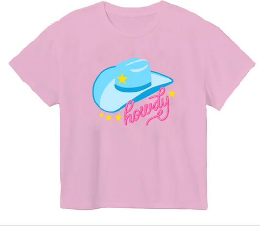 The Azarhia Rodeo Glitter Howdy Hat t-shirt is light pink and features a large blue rodeo hat with a yellow star, the word "howdy" in pink script, and small yellow stars surrounding the design.