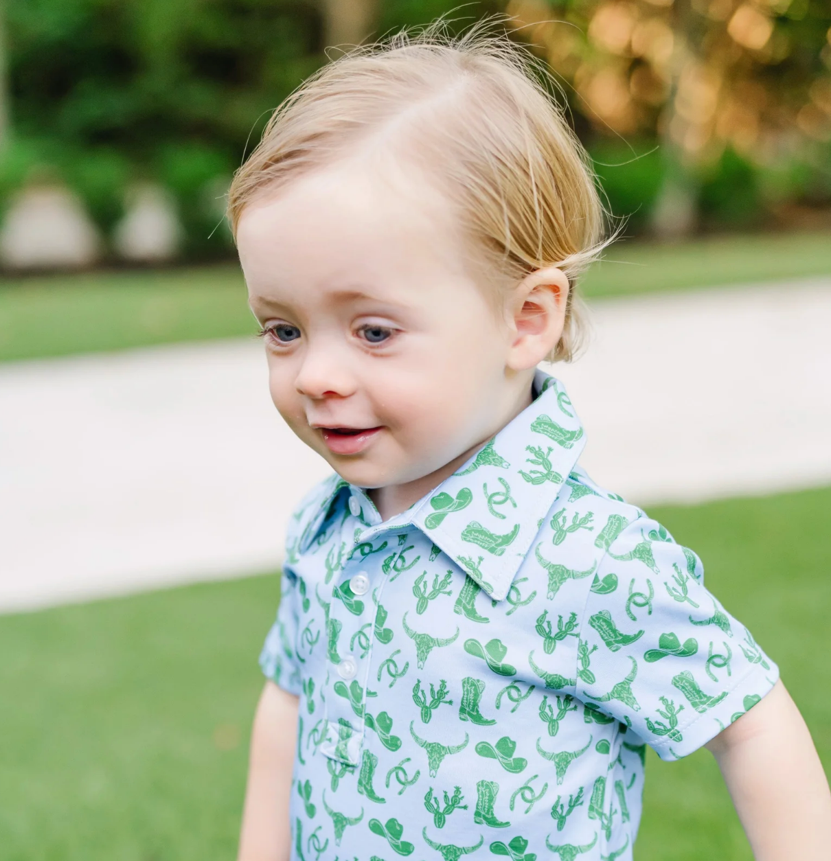 A young child with light hair is outdoors, wearing the Lila and Hayes Wildwest Tripp Shortall featuring green cactus designs. Blurred greenery and a white path appear in the background.