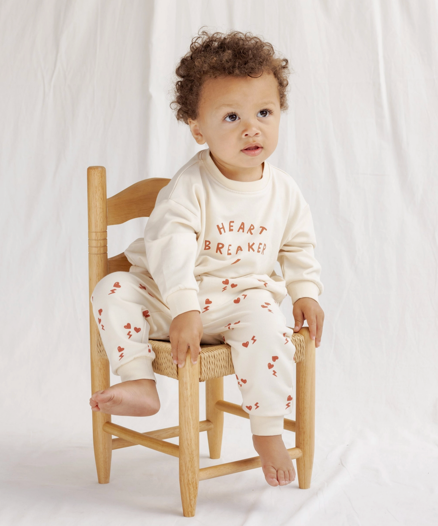 A toddler with curly hair sits barefoot on a wooden chair, wearing Quincy Mae's Electric Heart Relaxed Sweatpant and matching sweatshirt, both white with red hearts, standing out against a plain white fabric background.