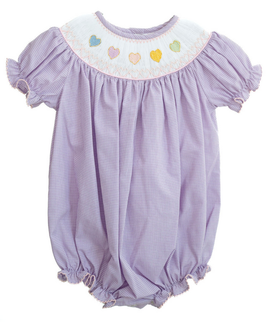 The Ruth & Ralph Rainbow Hearts Birdie Bubble is a lavender baby romper with short puffed sleeves, gathered hems, bishop smocking, and a collar adorned with pastel rainbow heart embroidery.