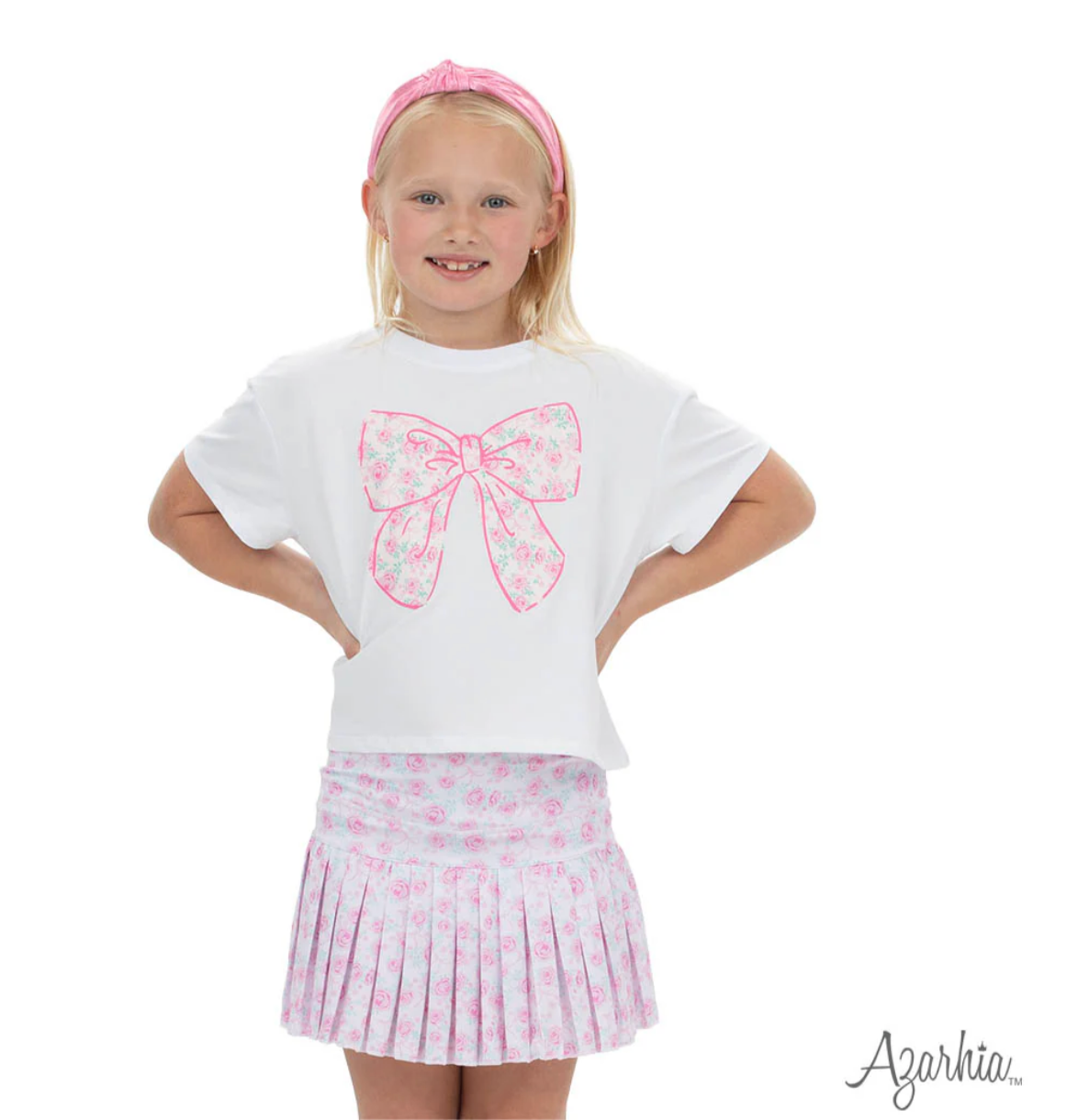 A young girl smiles, wearing the Azarhia Big Bow in Rosie Print T featuring a bold floral bow, paired with a matching pink pleated skirt and headband, standing hands on hips against a white background.