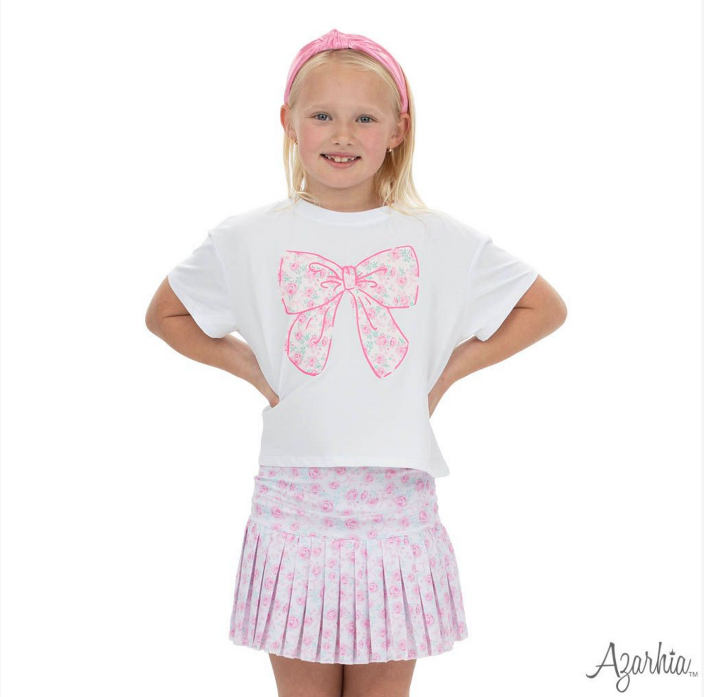 A young girl smiles in a white shirt with a large pink floral bow graphic, pink headband, and the Azarhia Rosie Bows Mini Pleat Skort, standing hands on hips against a white background.