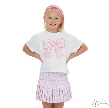 A young girl smiles in a white shirt with a large pink floral bow graphic, pink headband, and the Azarhia Rosie Bows Mini Pleat Skort, standing hands on hips against a white background.
