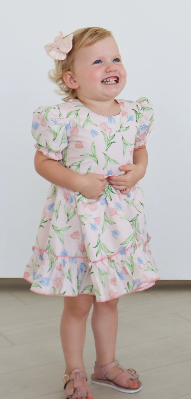 A young child with blond hair smiles indoors, hands clasped, wearing a Hollis Baker Everly Tulips Dress and pink sandals.