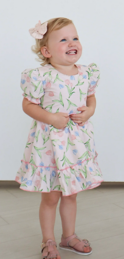 A young child with blond hair smiles indoors, hands clasped, wearing a Hollis Baker Everly Tulips Dress and pink sandals.