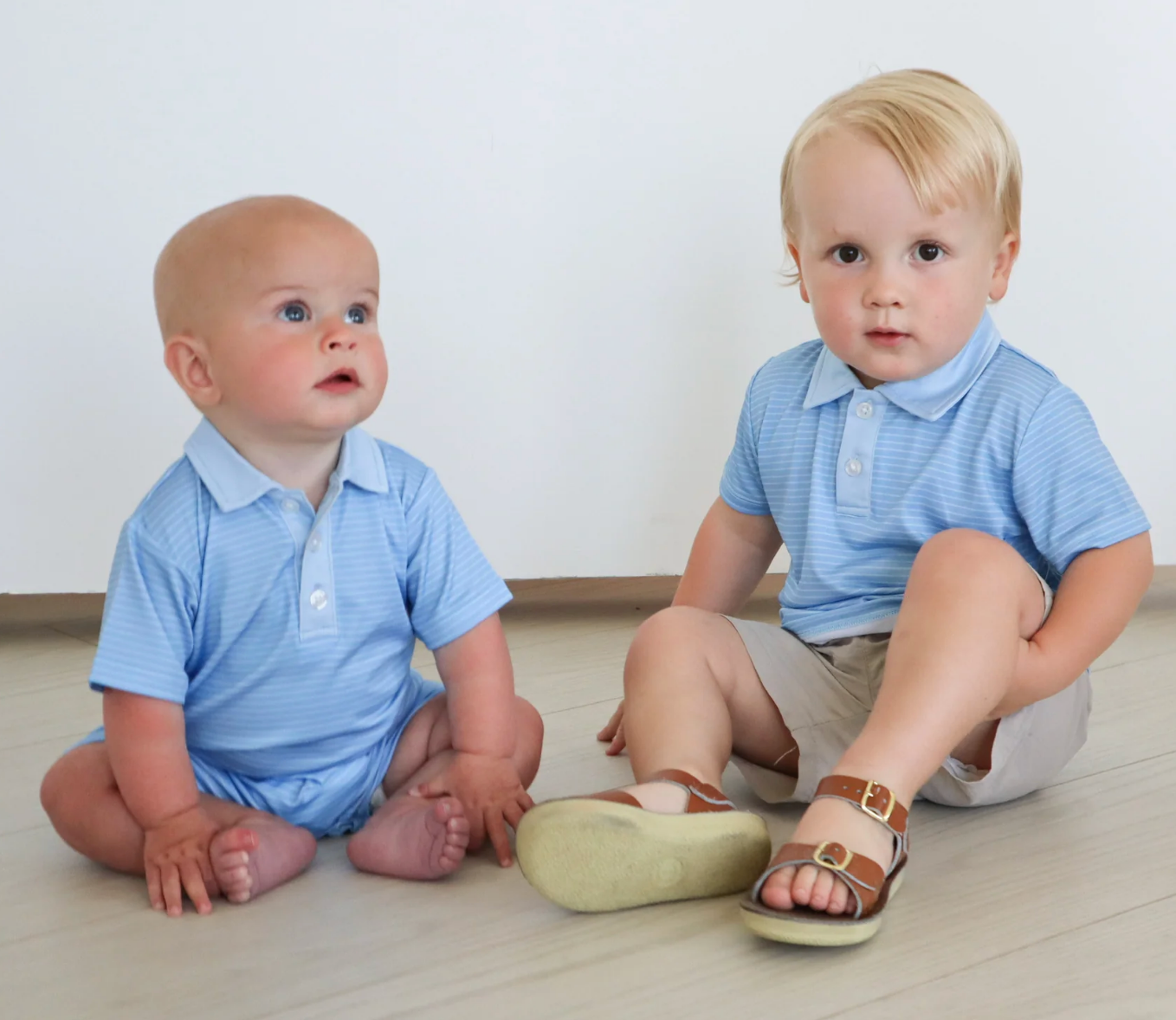Two young children sit on a light floor. The older wears a Hollis Baker Blue Stripe Try Polo, beige shorts, and sandals, while the baby is barefoot in a onesie. Both are in front of a white wall and wearing light blue collared shirts.
