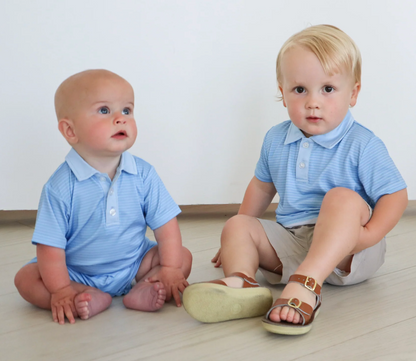 Two young children sit on a light floor. The older wears a Hollis Baker Blue Stripe Try Polo, beige shorts, and sandals, while the baby is barefoot in a onesie. Both are in front of a white wall and wearing light blue collared shirts.