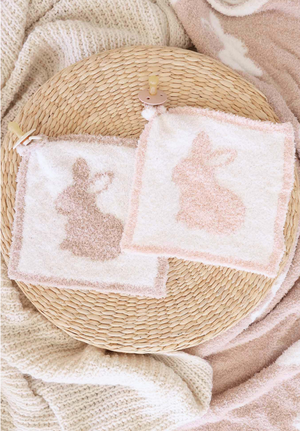 The CuddleLane™ Luxe Lovey | Bunny Foo Foo by Caden Lane features two square washcloths with bunny silhouettes in light brown and peach on a woven surface, surrounded by soft neutral blankets—perfect for added baby comfort.
