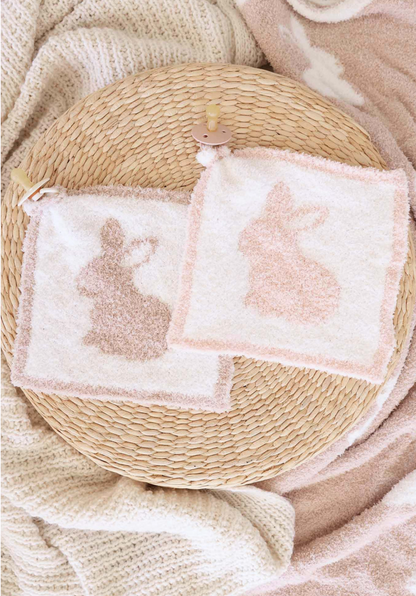 The CuddleLane™ Luxe Lovey | Bunny Foo Foo by Caden Lane features two square washcloths with bunny silhouettes in light brown and peach on a woven surface, surrounded by soft neutral blankets—perfect for added baby comfort.