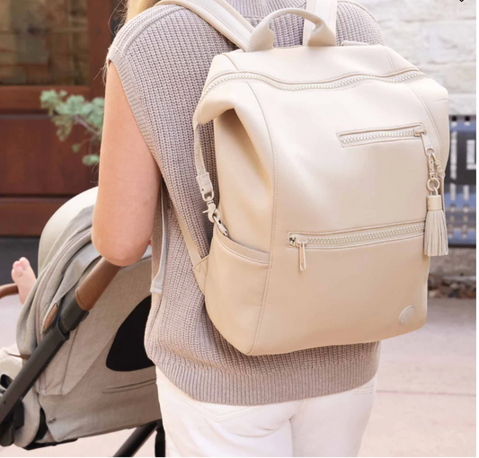 A person with long hair, wearing a beige sleeveless top, carries the Caden Lane Weslie Diaper Backpack Sand—featuring an insulated bottle holder—while pushing a stroller outdoors in a casual, everyday setting.