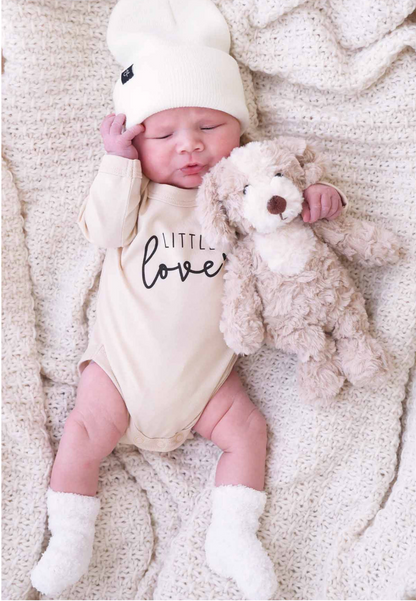 A baby in a white hat, cream "little love" onesie, and white socks lies on a textured blanket holding the Caden Lane Cuddle Plush Friends—an adorable plush brown and white dog, perfect as a baby gift.