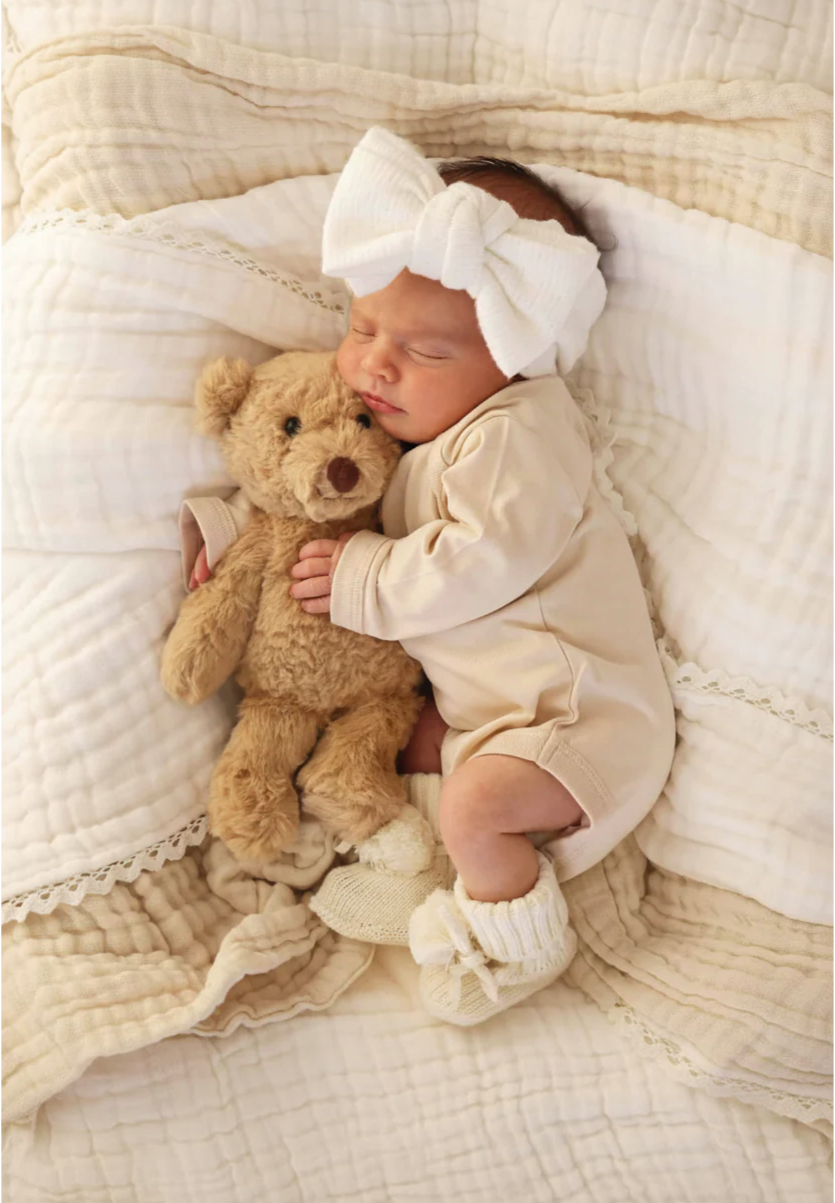A sleeping baby in a cream outfit and large white headband cuddles the Caden Lane Cuddle Plush Friends teddy bear while resting on a quilted blanket—a heartwarming scene and perfect baby gift.