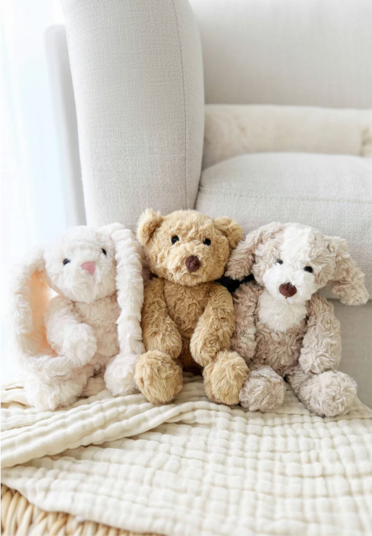 The Cuddle Plush Friends by Caden Lane—a white bunny, brown teddy bear, and beige dog—rest on a cream blanket, making them the perfect cozy baby gift or newborn companions in front of a light sofa.