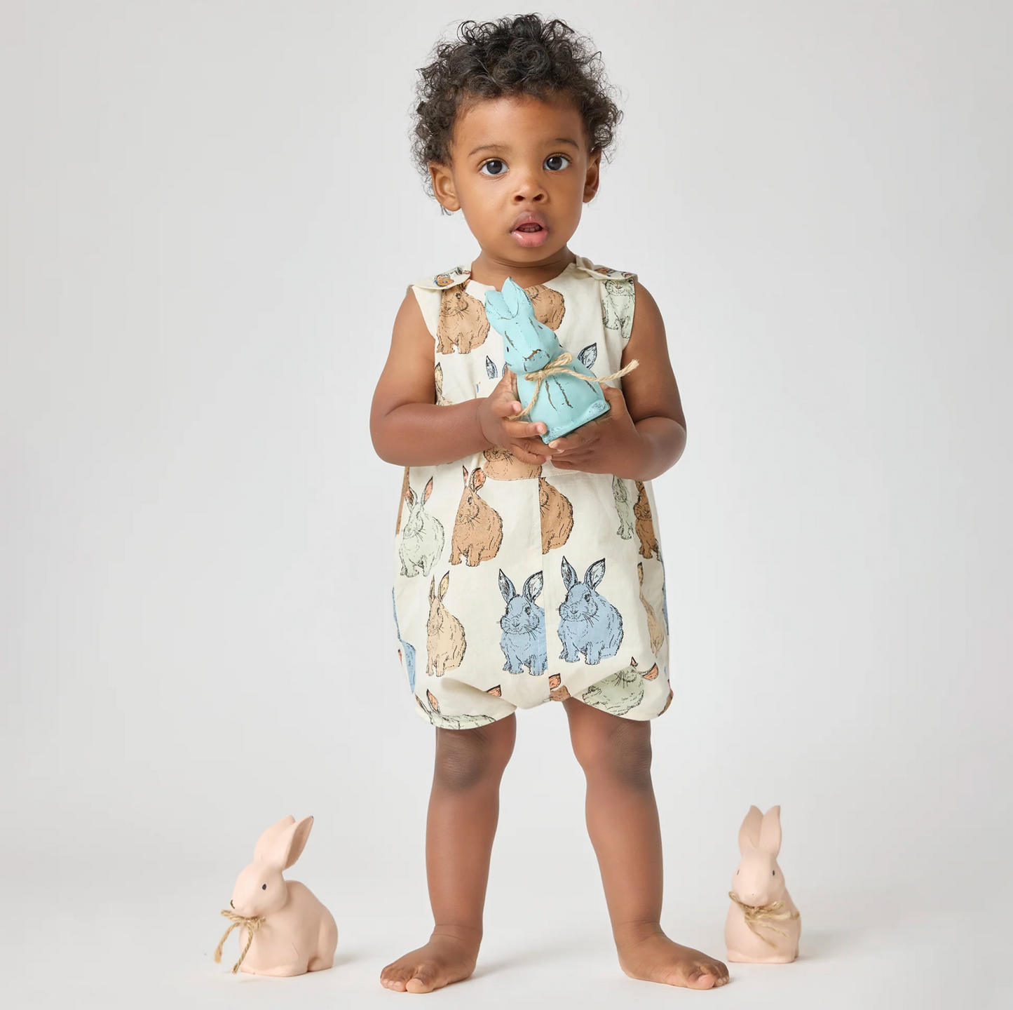 A young child stands barefoot on a light background, dressed in the Pink Chicken Vintage Bunny Noah Jumper and holding a blue stuffed rabbit. Two ceramic bunny figurines with gold ribbons complete this charming Easter scene.
