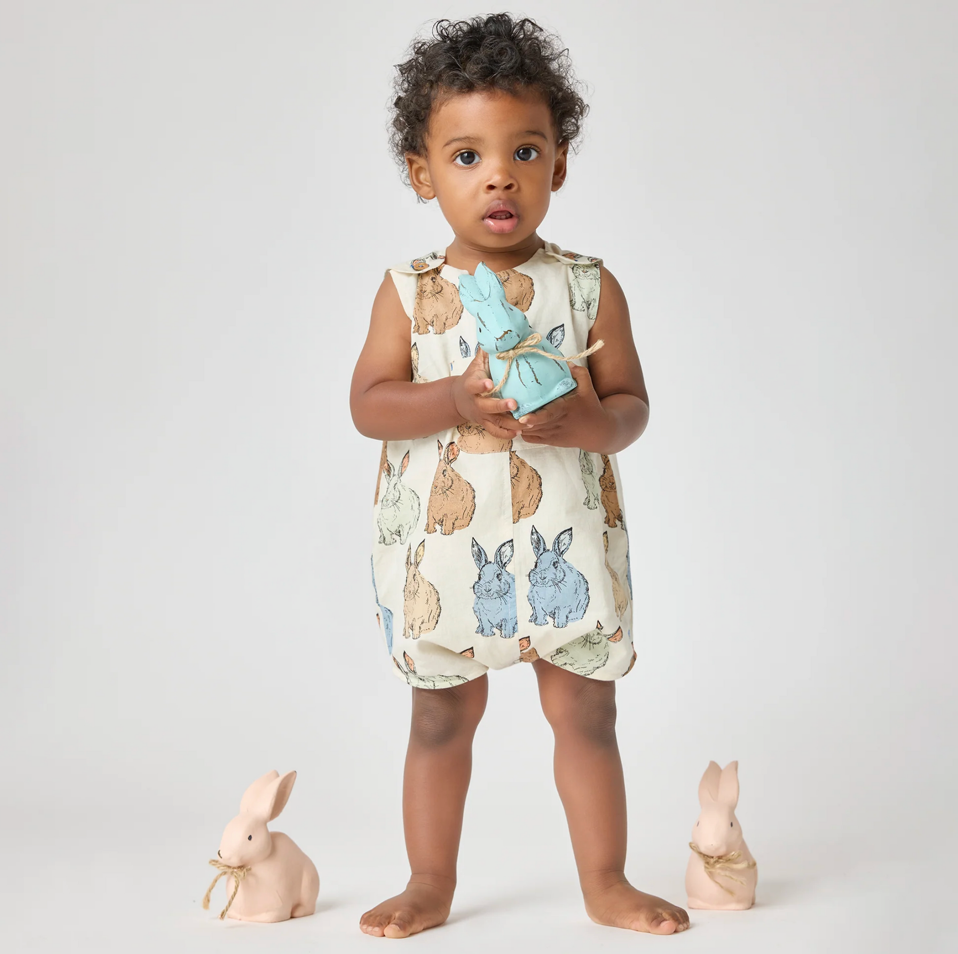 A young child stands barefoot on a light background, dressed in the Pink Chicken Vintage Bunny Noah Jumper and holding a blue stuffed rabbit. Two ceramic bunny figurines with gold ribbons complete this charming Easter scene.