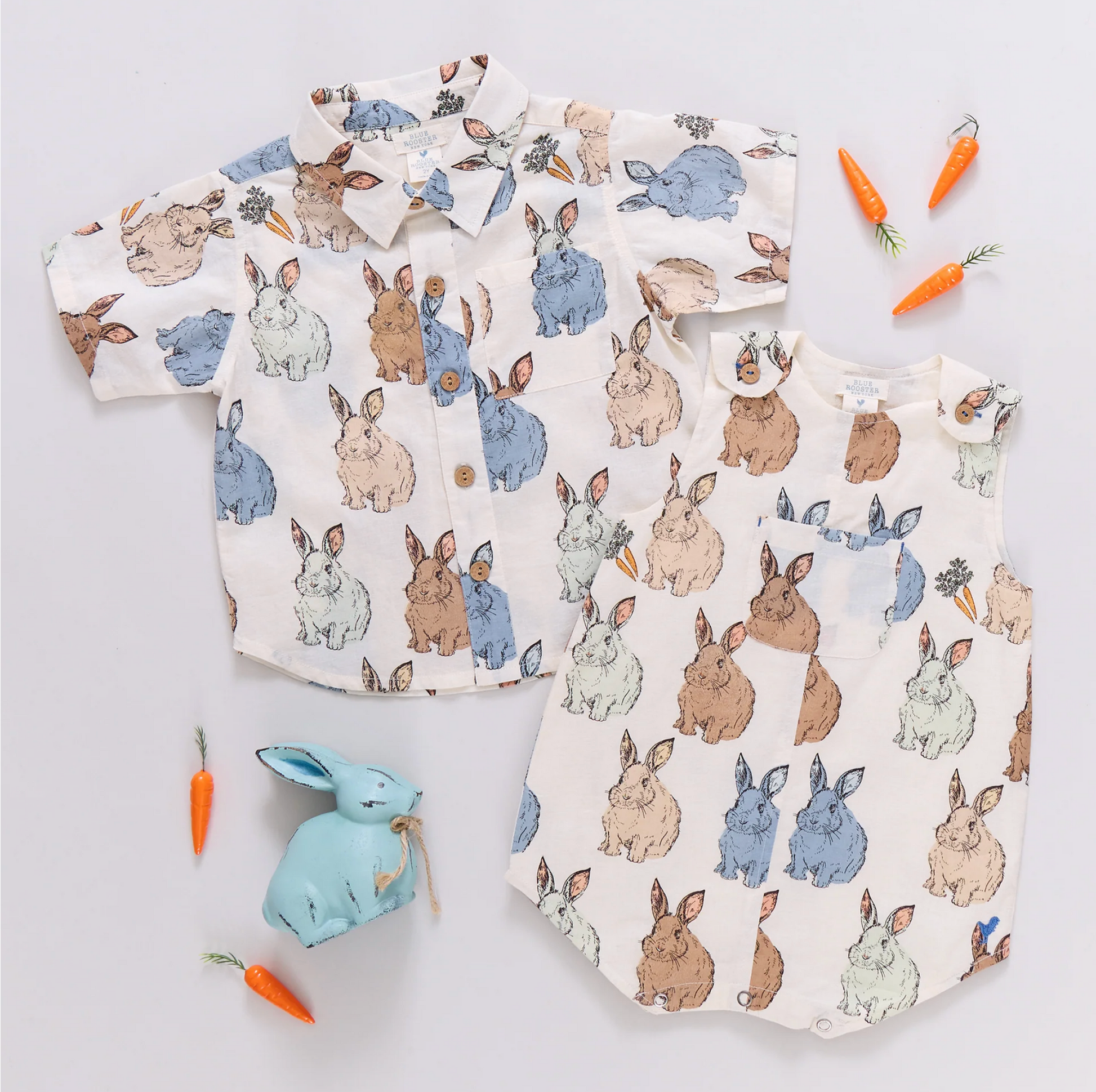 The Pink Chicken Vintage Bunny Noah Jumper, a sleeveless romper with cheerful bunny prints, is displayed alongside toy carrots and a small blue stuffed bunny on a white background.