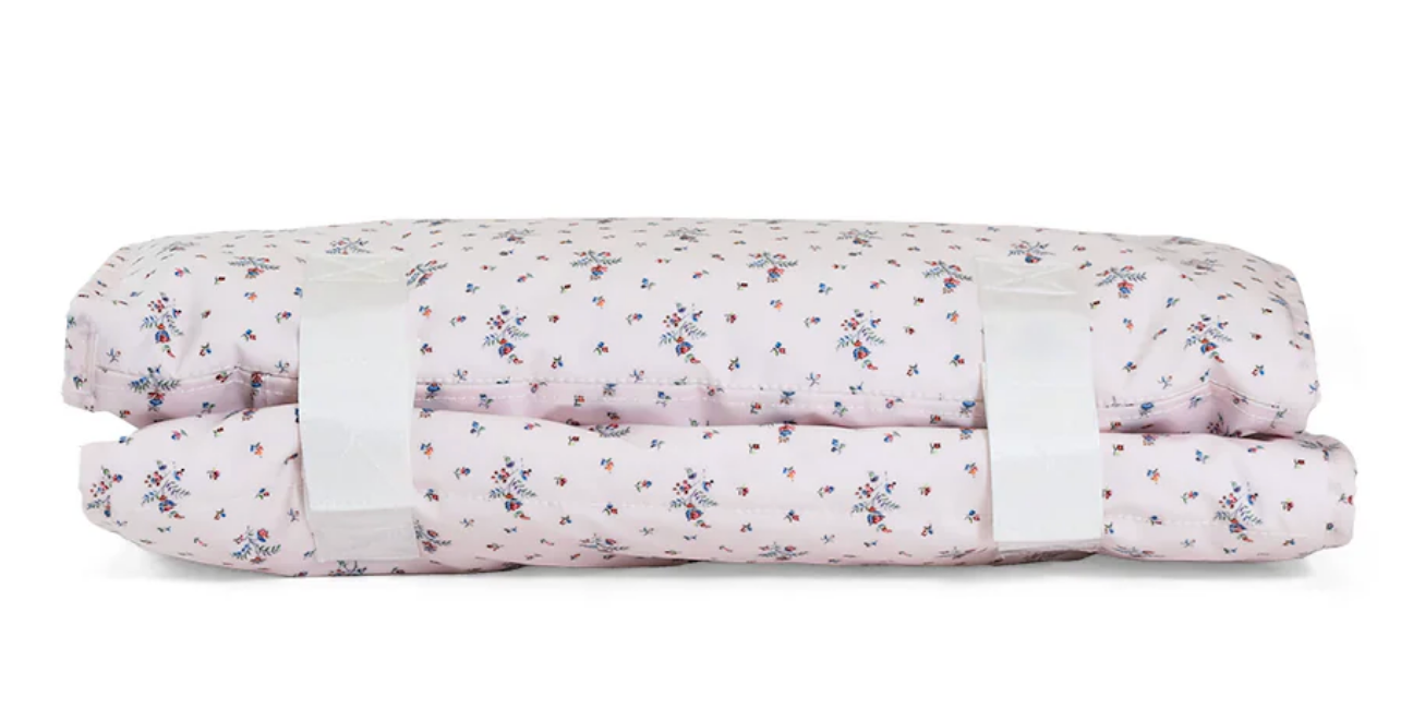 The TRVL Nap Mat by TRVL is a folded, light pink mat with a small floral pattern and two white straps for securing; it's machine washable for easy cleaning.