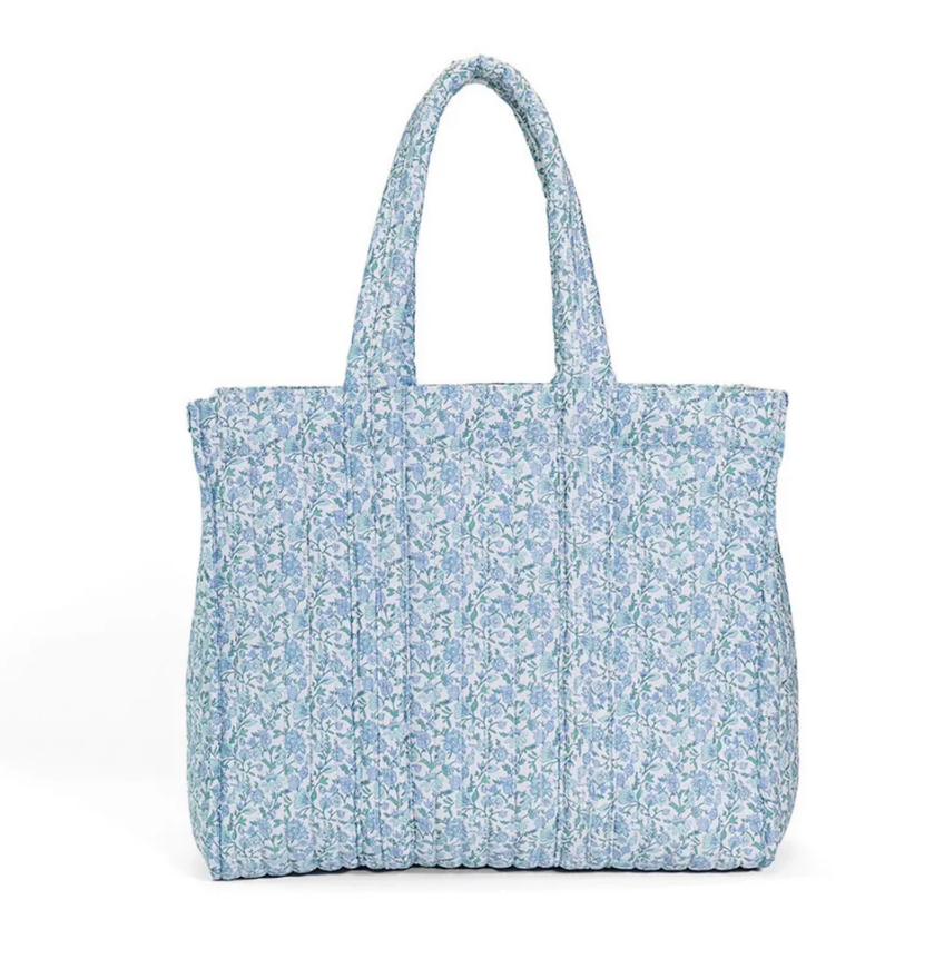 The TRVL QUILTED GO GO TOTE is a light blue and white floral quilted tote bag with padded handles. Machine washable, it stands upright against a plain white background.