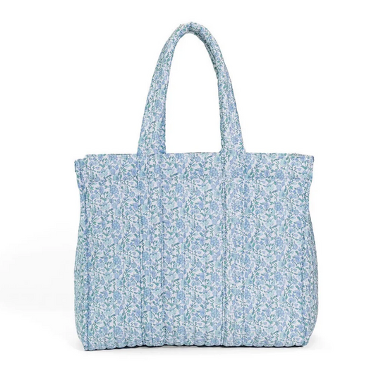 The TRVL QUILTED GO GO TOTE is a light blue and white floral quilted tote bag with padded handles. Machine washable, it stands upright against a plain white background.
