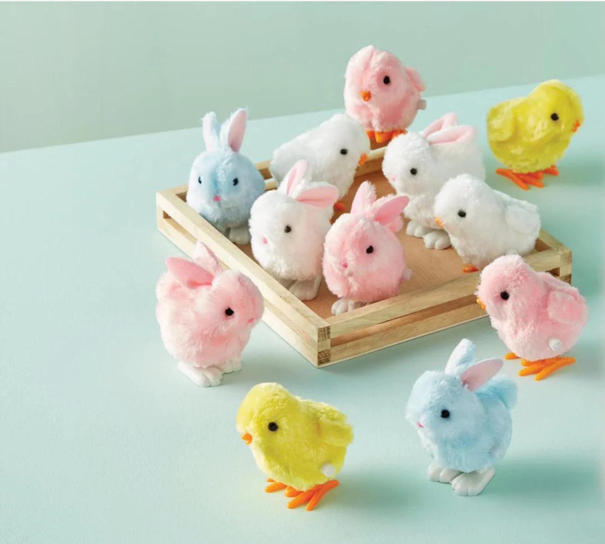 The mudpie Wind Up Chicks and Bunnies—fluffy yellow, pink, blue, and white Easter basket toys—are arranged around and inside a wooden tray on a light blue surface.