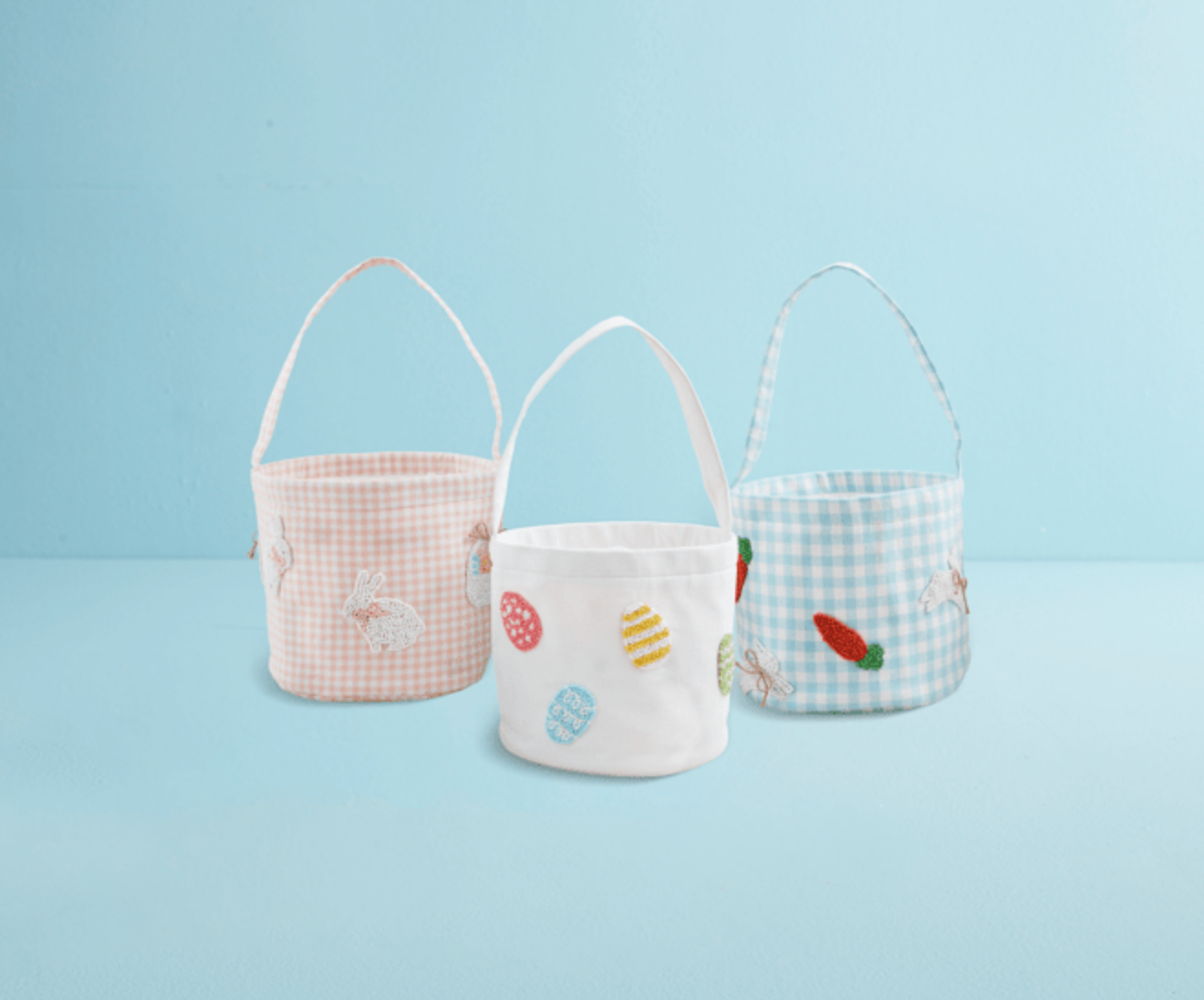 Three mudpie Beaded Easter Buckets—pink plaid with bunnies, white with colorful eggs, and blue plaid cotton with carrot and bunny designs—are displayed side by side on a light blue background.