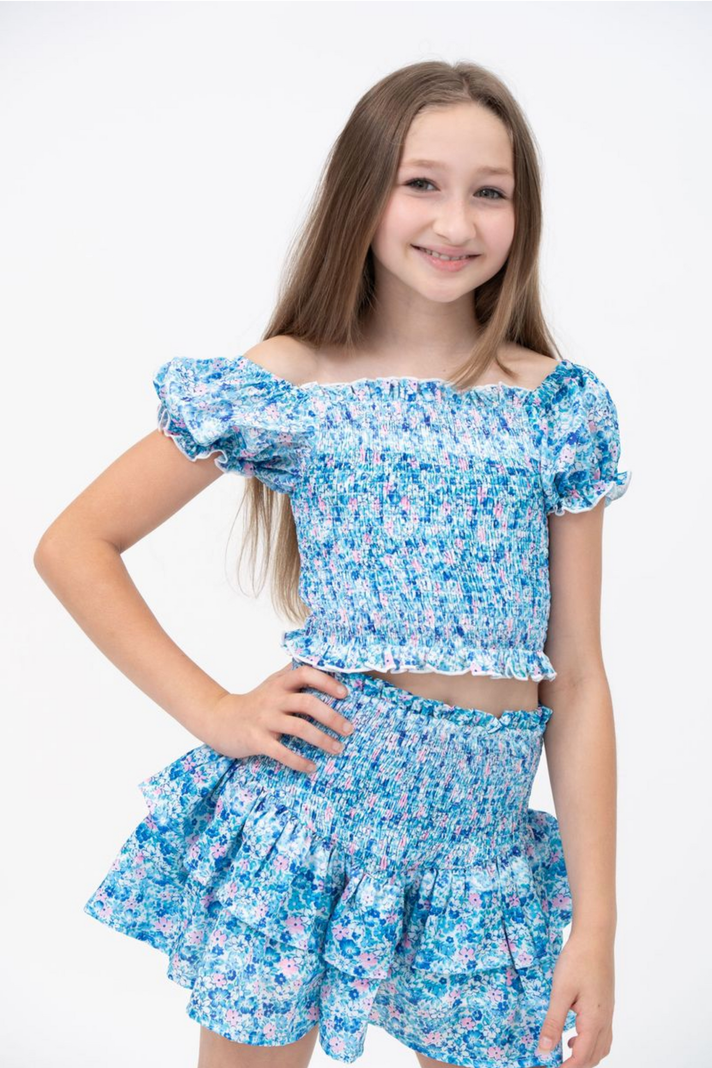 A young girl with long brown hair smiles in the Tweenstyle by Stoopher Blue & Pink Ditzy's Print Smocked Set, featuring an off-the-shoulder top and matching ruffled skirt, perfect for girls’ clothing, against a plain white background.