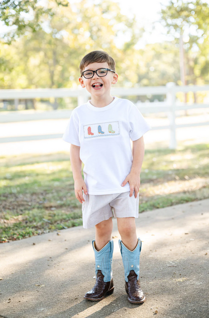 A young boy stands smiling outside in the Ruth & Ralph Cowboy Boots Smocked Beau T-Shirt/Shorts Set, bringing southern charm to the scene with his gingham shorts and cowboy boot details, against a backdrop of trees and a white fence.