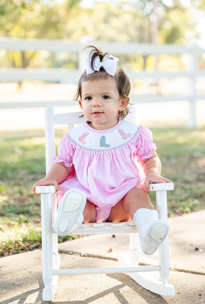 A toddler with brown hair and a white bow sits on a small white rocking chair outside, wearing the Ruth & Ralph Cowgirl Boots Smocked Birdie Bubble. The scene features green grass, trees, and a white fence in the background.