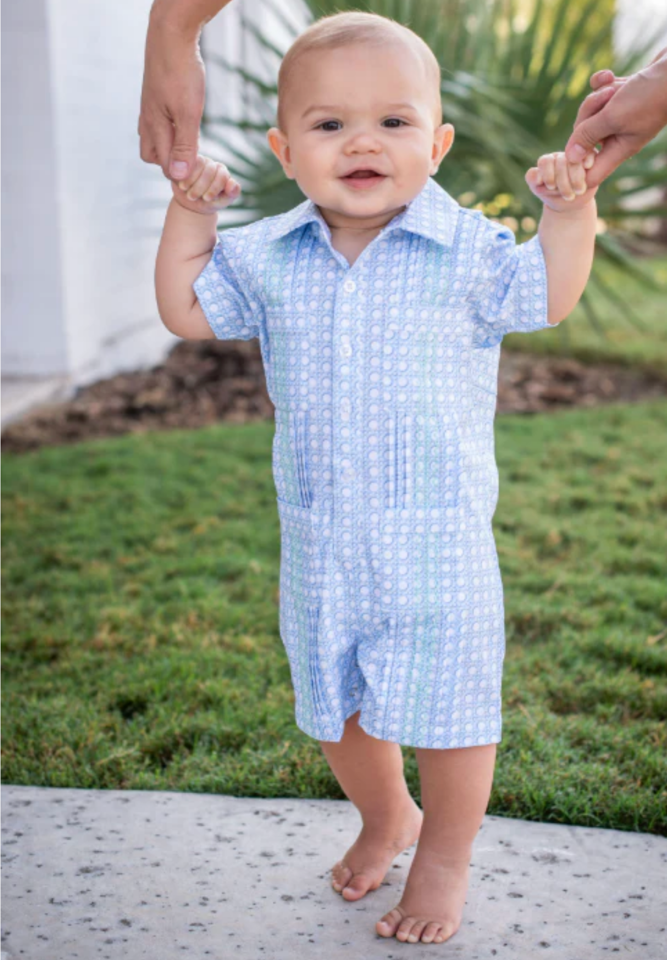 A smiling baby in the blue Quail Blue Cane Lattice Guayabera Romper, featuring UPF 50+ sun protection, stands on a sidewalk holding hands with two adults, surrounded by grass and plants.
